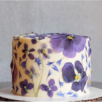 A cake frosted with smooth, white buttercream and decorated all over with purple pressed flowers.