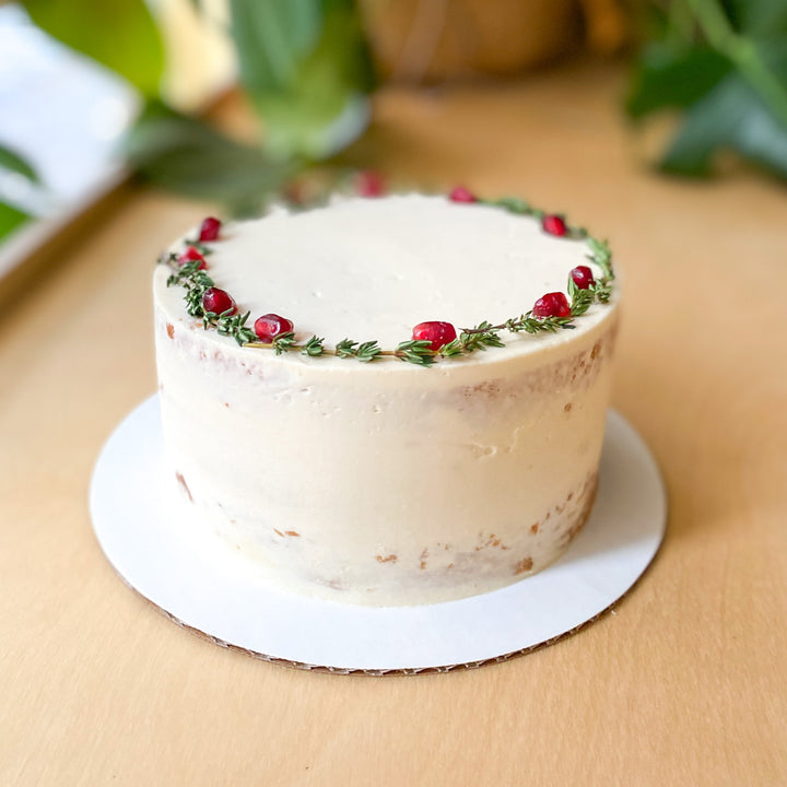 A round cake covered in a sheer layer of white buttercream and decorated with a crown of greenery and pomegranate seeds.