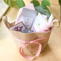 A small burlap tote containing two pouches of loose-leaf tea, a small notecard, and a jar of sugar cubes. The tote is tied with a pink ribbon.
