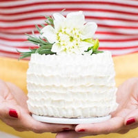 A woman holding a miniature cake decorated with white buttercream ruffles and topped a fresh flower.