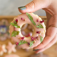 A hand holding a wreath-shaped cookie decorated with colored glaze and pressed florals.