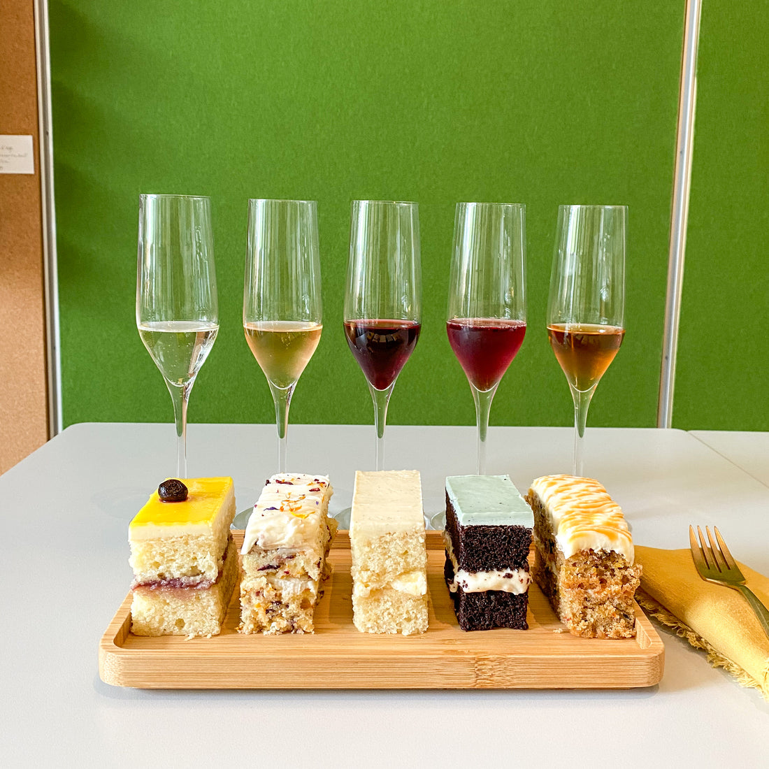 A platter with five rectangular slices of cake, each a different flavor, placed beside five flutes of wine containing 1 ounce tasting portions.
