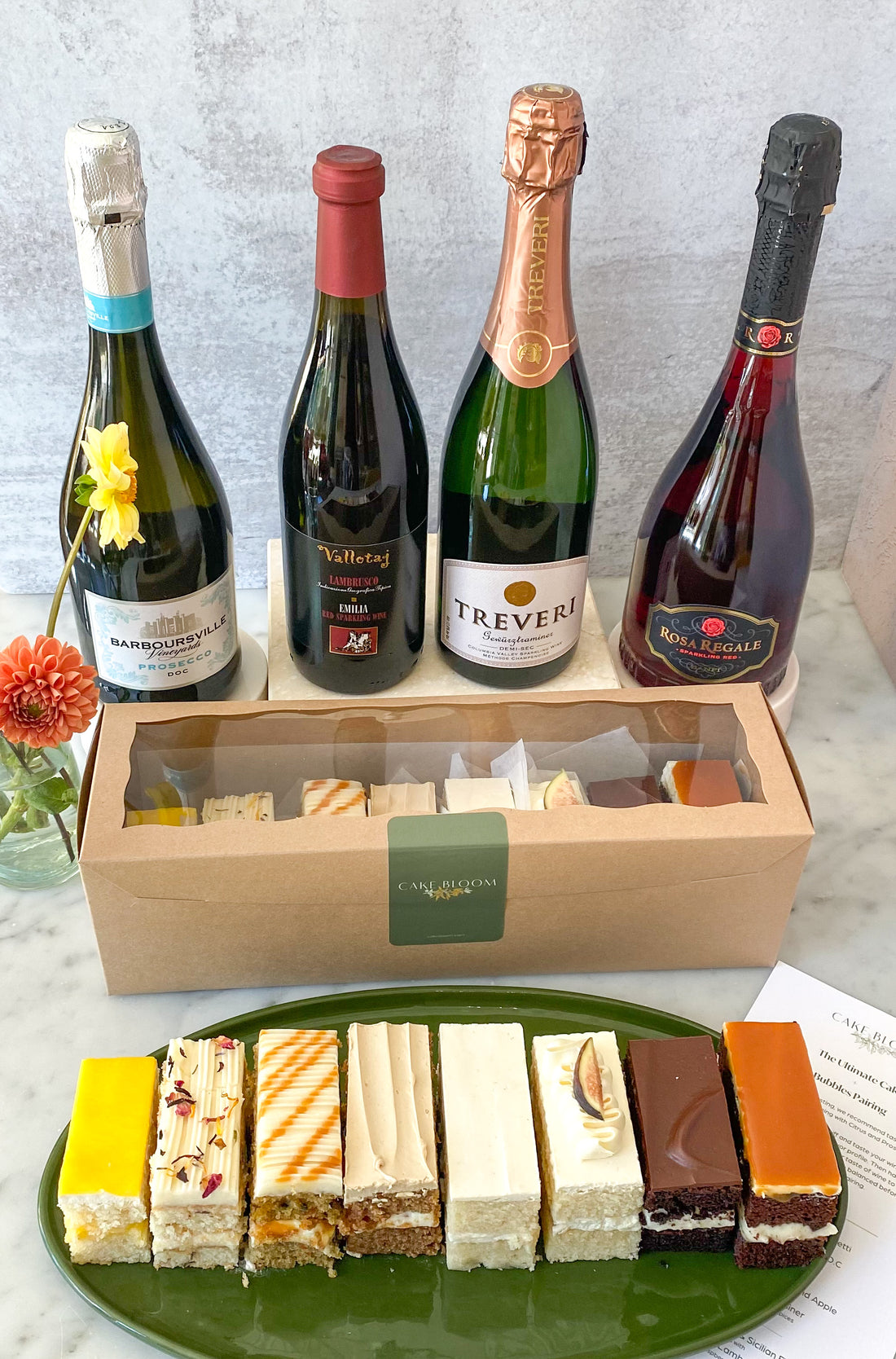 A ceramic platter with eight slices of cake, each a different flavor, beside a box filled with the same. Four bottles of wine are displayed behind the cake.