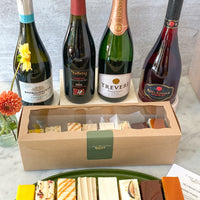 A ceramic platter with eight slices of cake, each a different flavor, beside a box filled with the same. Four bottles of wine are displayed behind the cake.