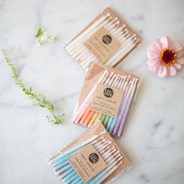 Three packs of candles laid on a table; one white, one ombre blue, and one ombre rainbow.