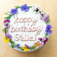 A cake decorated with a flower crown and the words "happy birthday stella!" in pink cursive buttercream