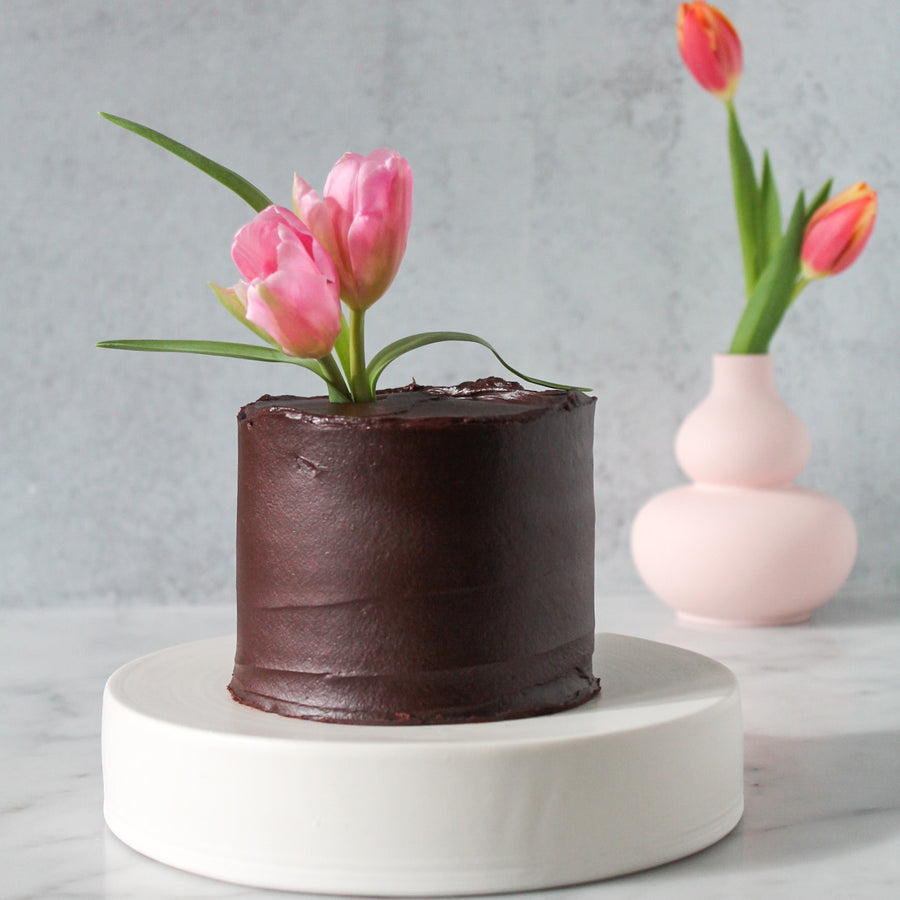 A small round cake covered in ganache and topped with a bouquet of pink tulips.