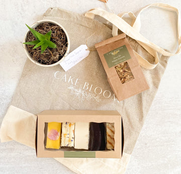 A tote bag printed with the Cake Bloom logo, laid out on a table. On top of the bag are a succulent, a bag of granola, and a box of cake slices. 