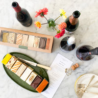 A ceramic platter with eight slices of cake, each a different flavor, beside a box filled with the same. Four bottles of wine sit beside a pile of plates and forks, as well as a printed tasting guide.