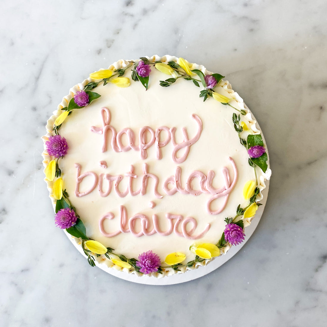 A cake decorated with a flower crown and the words "happy birthday claire" in pink cursive buttercream
