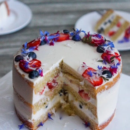A round cake covered in sheer white buttercream and decorated with blueberries, strawberries, and blue flowers. The cake is sliced, revealing strawberries and blueberries inside.