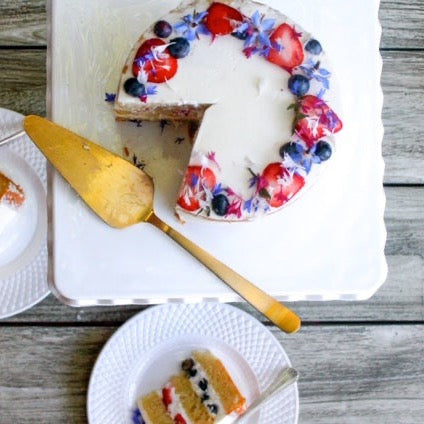 A round cake covered in sheer white buttercream and decorated with blueberries, strawberries, and blue flowers.